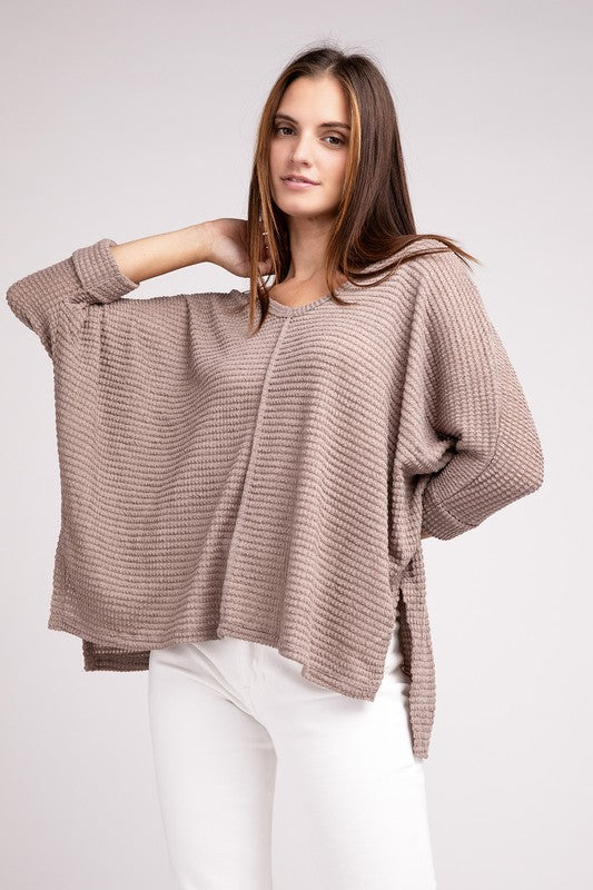 Model wearing a relaxed fit taupe sweater with a textured pattern and wide sleeves, styled with white pants.