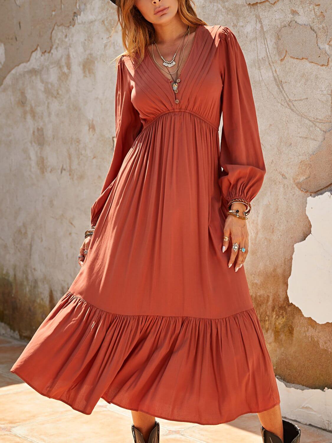 Woman wearing Ruched V-Neck Long Sleeve Midi Dress in rust color with ruffled detailing and accessorized with jewelry, standing against a rustic background.