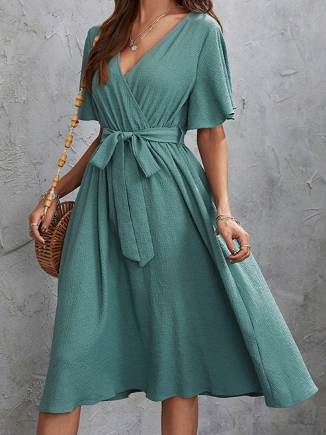 Woman wearing teal Surplice Flutter Sleeve Midi Dress with tie waist, holding a wicker bag, standing against textured grey background