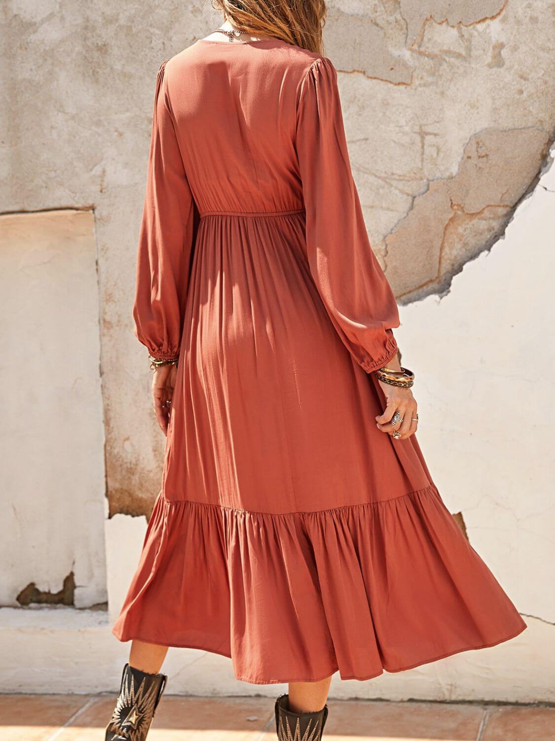 Woman wearing a rust-colored ruched V-neck long sleeve midi dress with ruffled details, standing outdoors against a rustic wall.