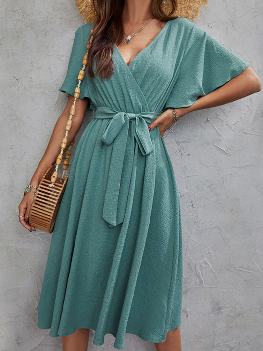 Woman wearing a teal Surplice Flutter Sleeve Midi Dress with a tied waist and carrying a woven handbag.