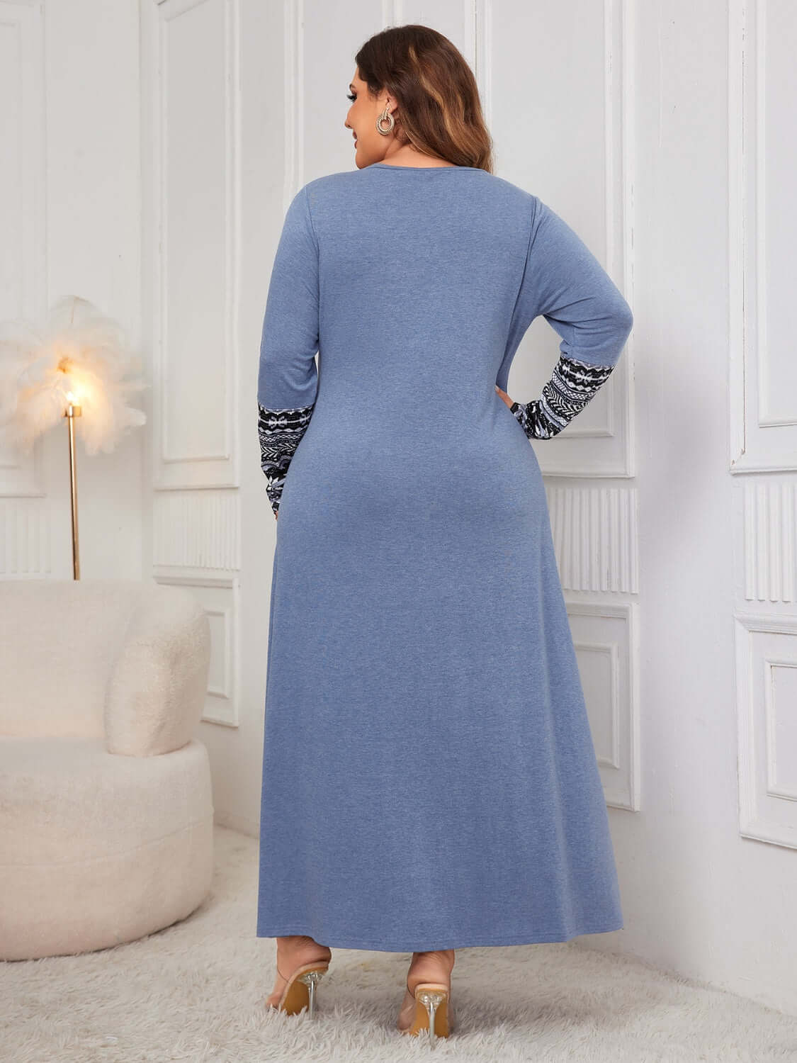 Plus size woman wearing a blue printed round neck long sleeve dress, view from the back, standing in a chic room with white walls.