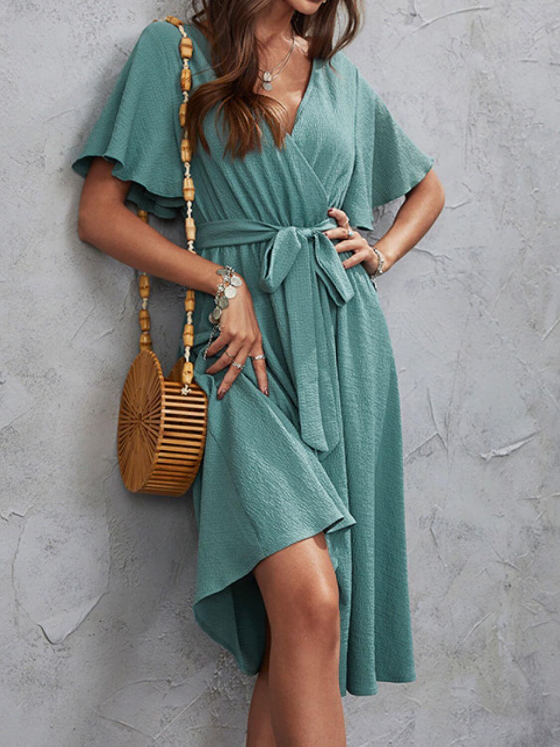 Woman wearing an elegant surplice flutter sleeve midi dress in teal, accessorized with a wooden handbag and jewelry, standing against a textured gray wall.