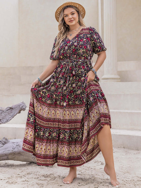 Plus size model in a printed V-neck short sleeve maxi dress with tied detail, standing on sandy steps wearing a hat.